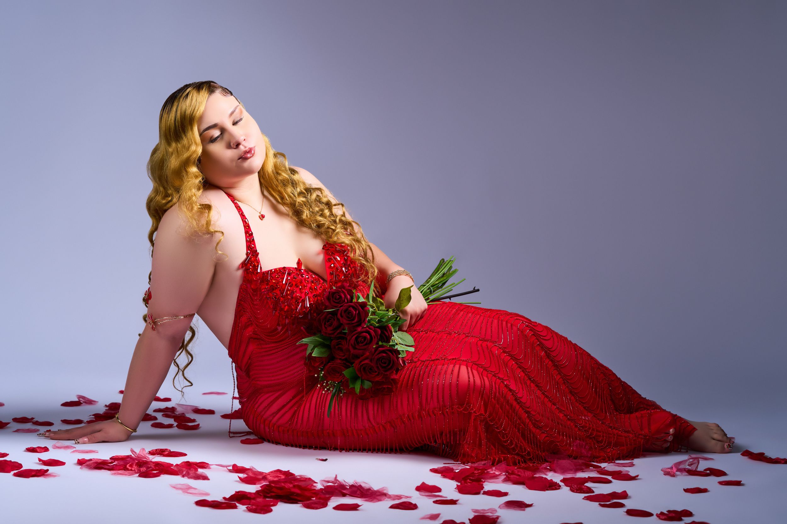 A person in a red evening gown poses with roses on white backdrop with scattered rose petals.