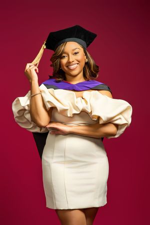 A graduate in academic regalia and white dress smiles while posing against a burgundy backdrop for a graduation portrait.