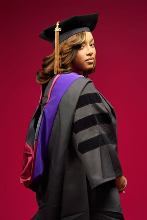 Graduate wearing black cap and gown with purple hood poses against burgundy background during commencement ceremony.