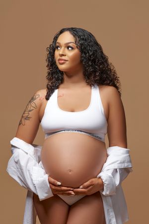 A pregnant woman in white sports bra and open shirt poses against brown background with arm tattoo visible.