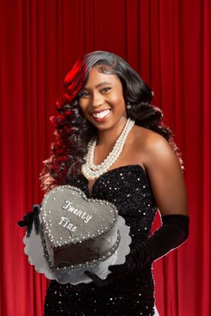 A glamorous portrait against a red curtain backdrop featuring sparkly black dress, long gloves and pearl necklace with heart dessert.