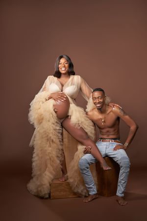 A maternity photoshoot features expecting parent in a tulle dress with wooden prop and partner in jeans against brown backdrop.