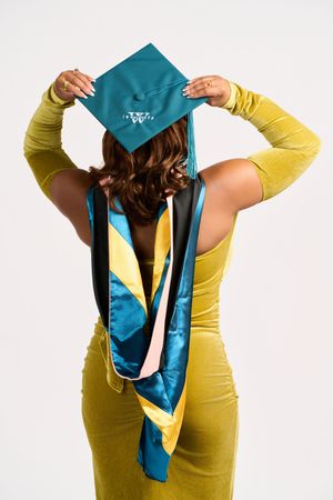 Graduate in yellow dress and teal graduation cap and stole poses with their back to the camera.