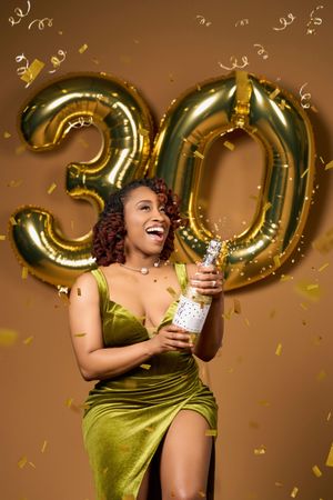 Woman in olive green dress laughs joyfully with gold number balloons and confetti in celebratory photo series.