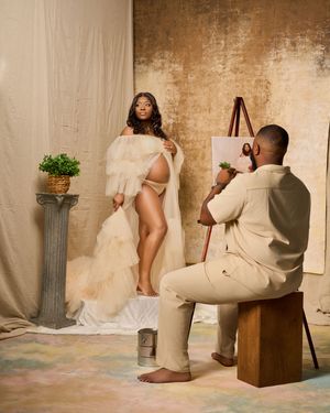 A photographer in beige clothing captures artistic photos with a camera while seated on a wooden block in a studio setting.