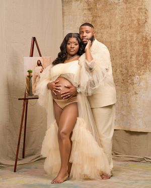 Elegant maternity photo featuring a couple in cream-colored outfits posing against a textured beige backdrop with an easel.