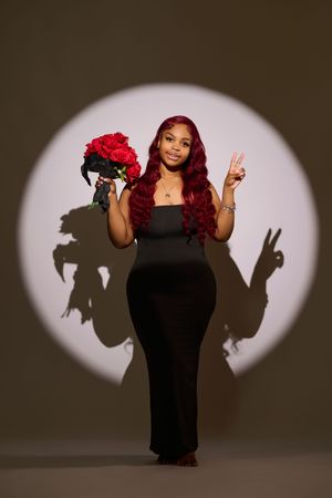 A figure in a black dress poses playfully with red roses against a neutral background creating dramatic shadows.