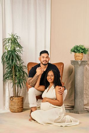 A couple sits together in a minimalist setting with indoor plants, wearing matching white outfits against a neutral backdrop.