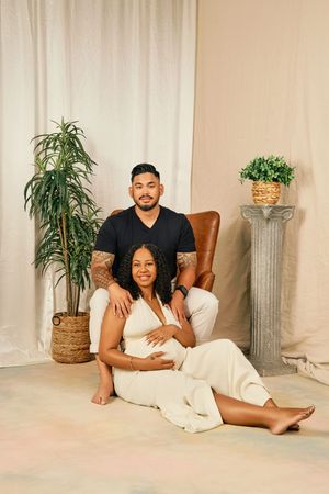 Maternity photo featuring couple in white and black outfits sitting on floor with plants and decorative column in background.