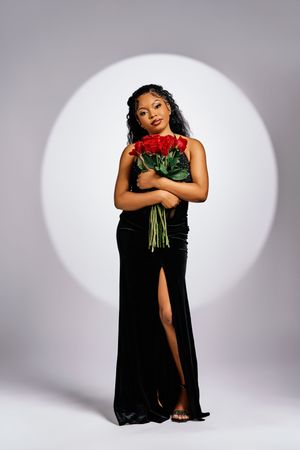 A figure in a black evening gown with thigh-high slit poses with red roses against a spotlight background.