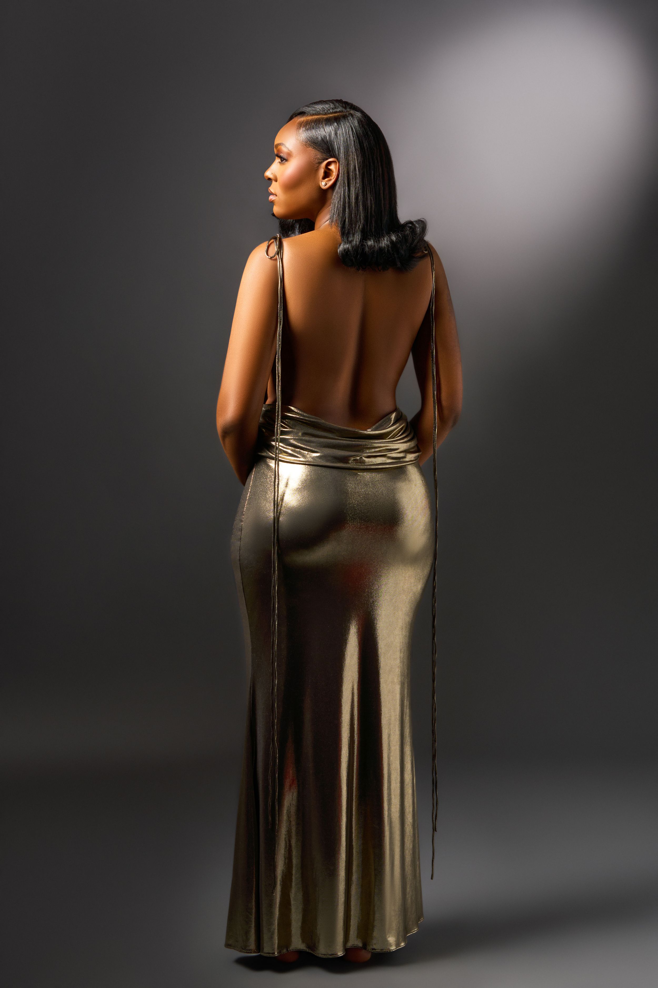 A person wearing a backless metallic gold evening gown poses against a dark background in a glamorous fashion photo.