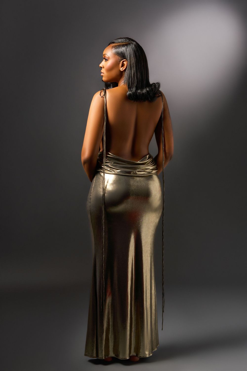 A person wearing a backless metallic gold evening gown poses against a dark background in a glamorous fashion photo.