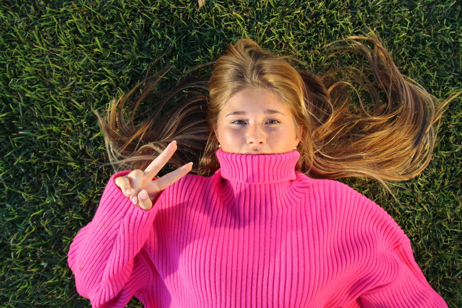 Person lying on grass wearing bright pink sweater making peace sign.