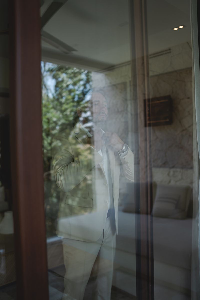 A ghostly reflection in a window pane shows a figure against green foliage and interior room elements.