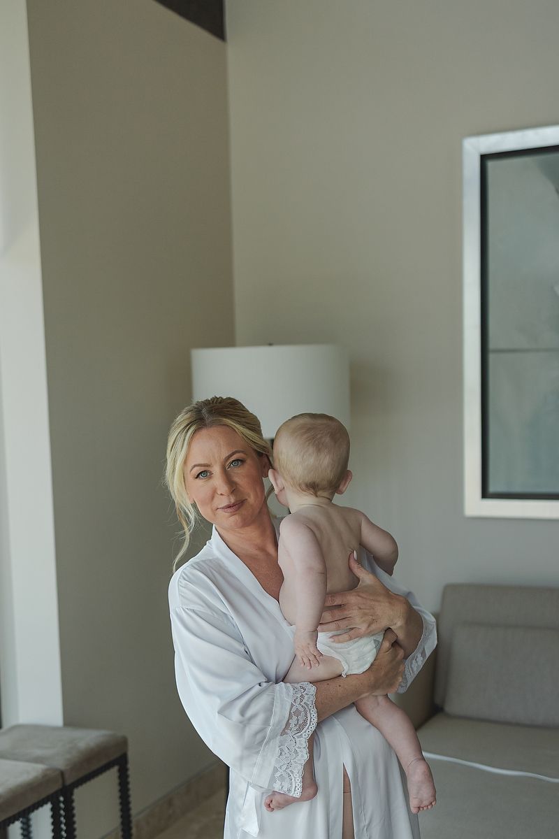 Woman in white robe holds baby in light-filled room with gray couch and white decor.