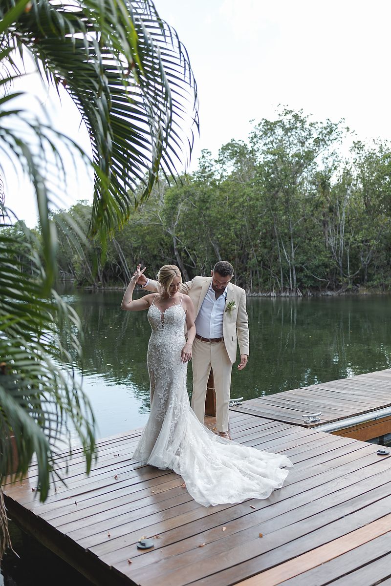 A couple in wedding attire stands on a wooden dock surrounded by tropical mangroves and palm trees near a tranquil lagoon.