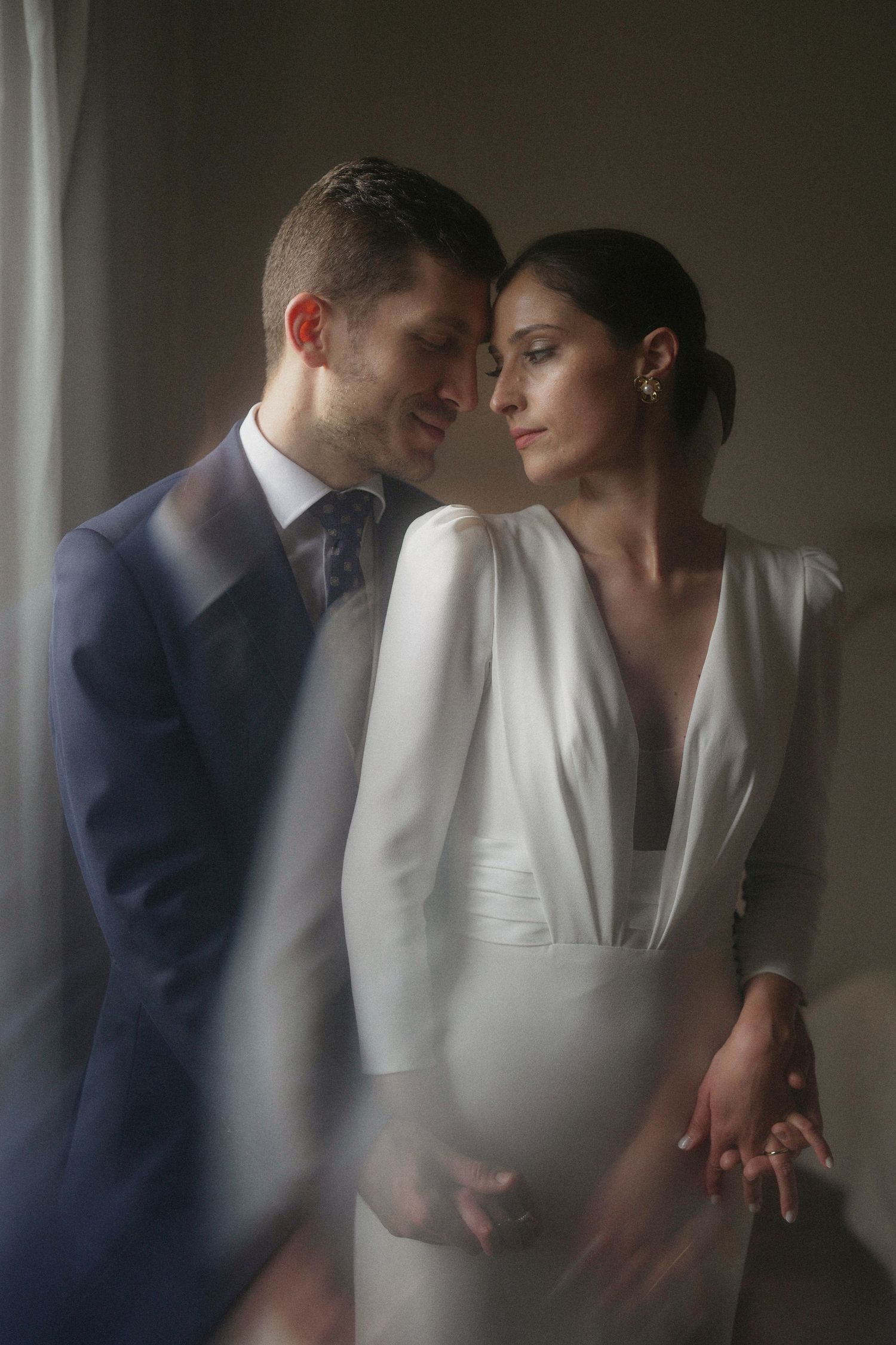 Intimate wedding portrait of couple in elegant white dress and navy suit standing together in soft window light.