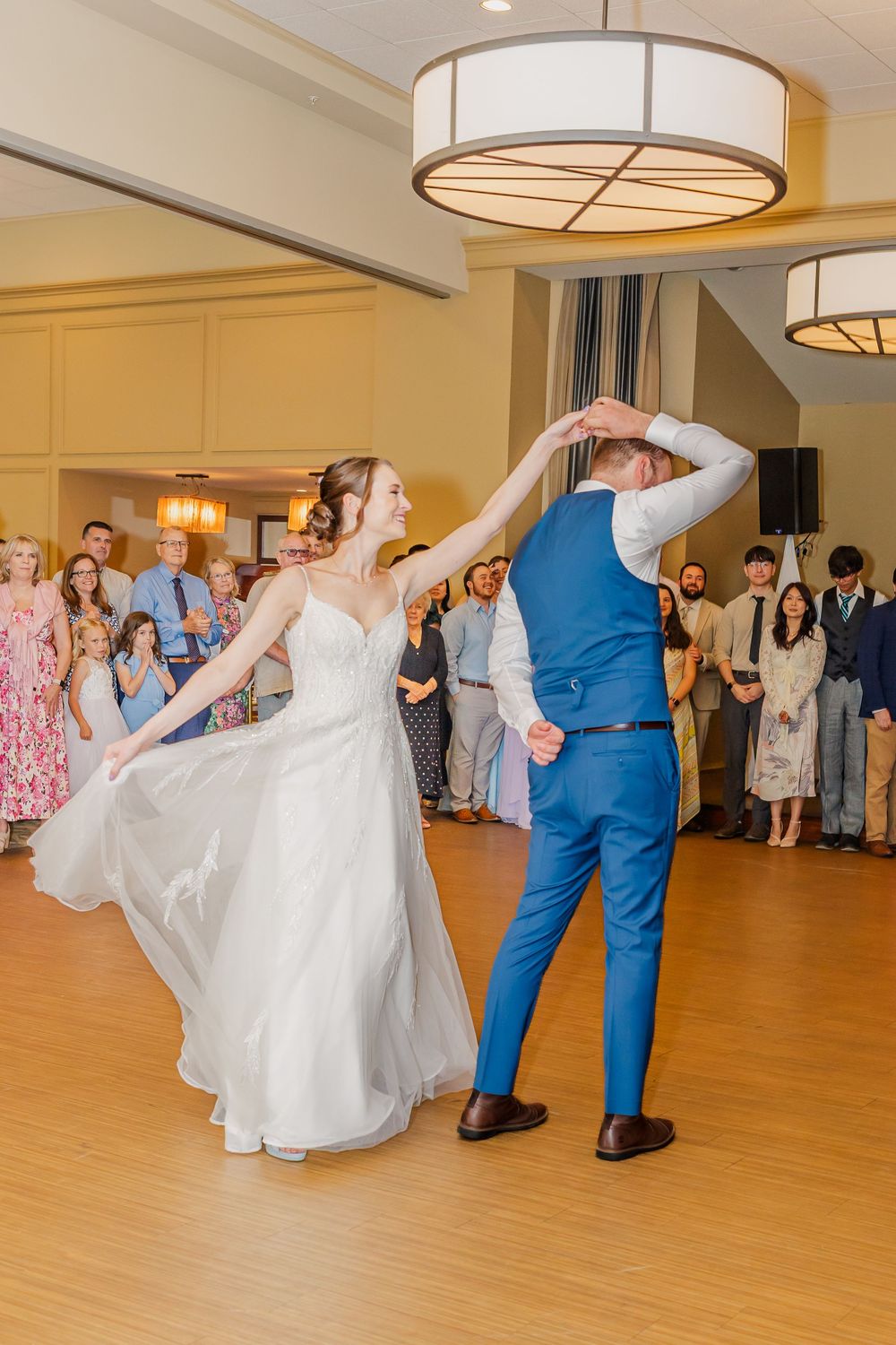 A couple share their first dance at a wedding reception as guests look on in a warmly lit ballroom.