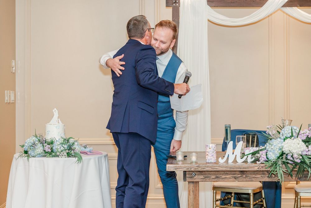 A wedding couple sharing an emotional first dance in an elegant reception hall decorated with white floral arrangements.