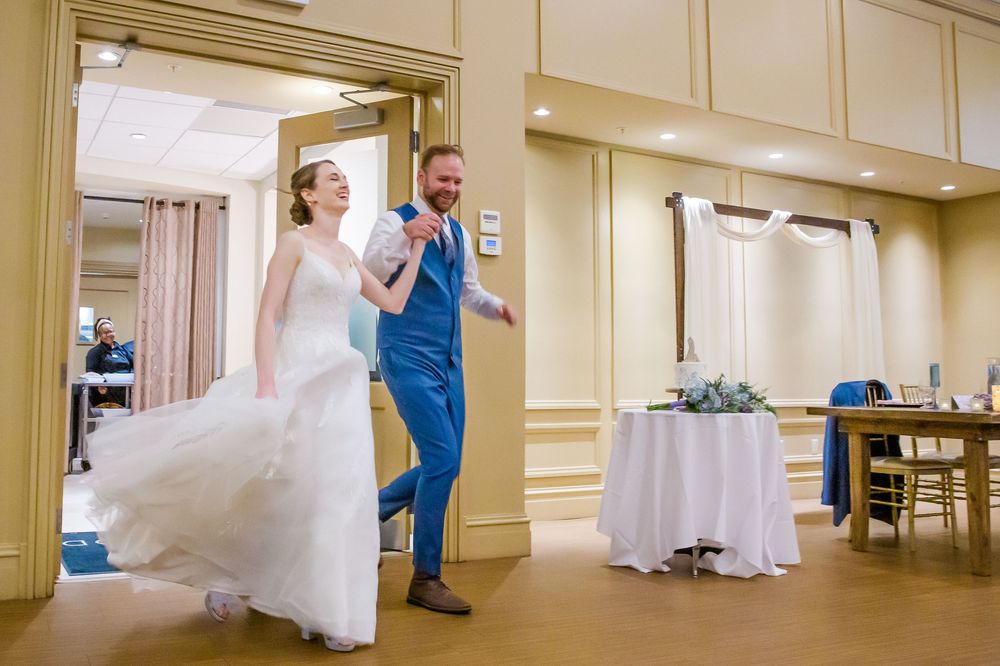 Two people prepare wedding dresses in a cream-colored hallway with ornate crown molding.