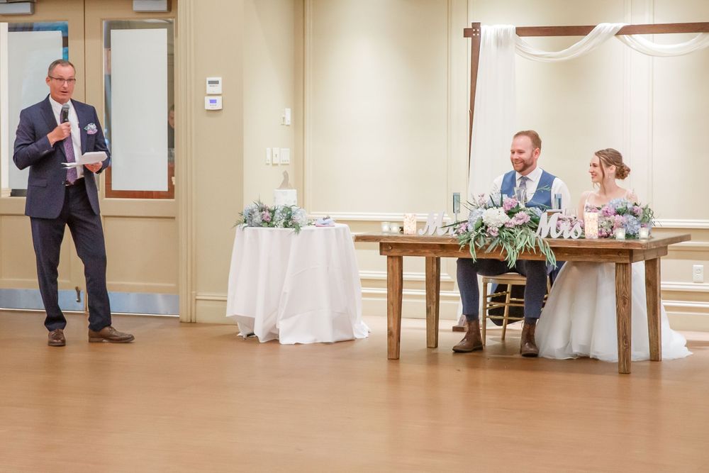 Wedding reception scene with people at a head table and someone delivering a speech in a banquet hall setting.