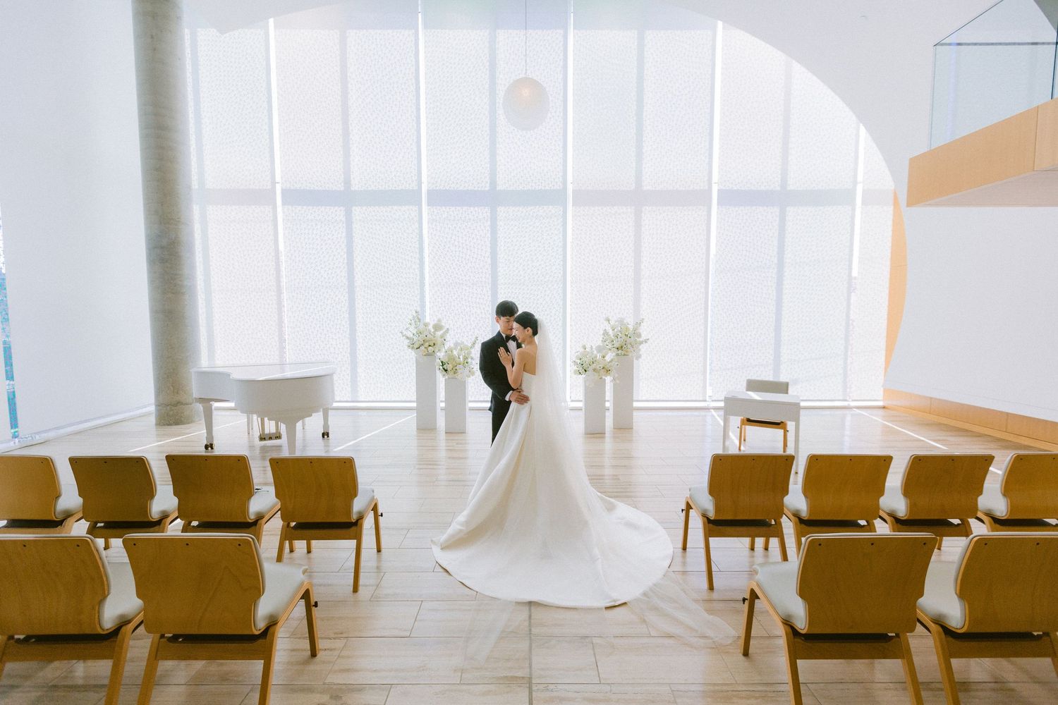 A minimalist white wedding ceremony setup with gold chairs and sheer white fabric draping in a bright modern space.