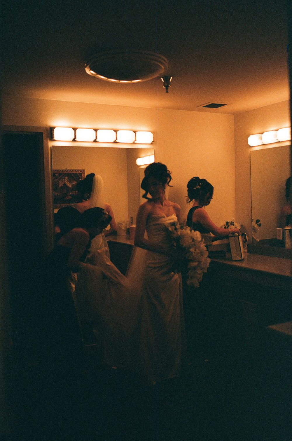 Film photo of bride in white gown preparing with bridesmaids in warmly lit dressing room mirror.