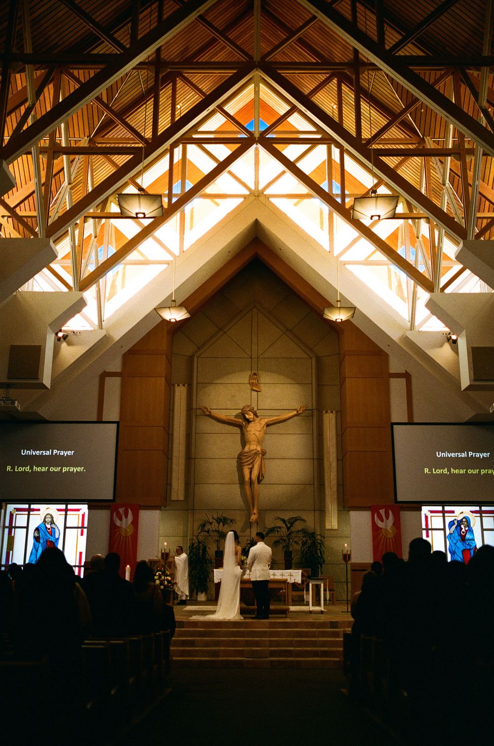 Wedding ceremony at Catholic church altar beneath large crucifix with Universal Prayer displayed on screens.