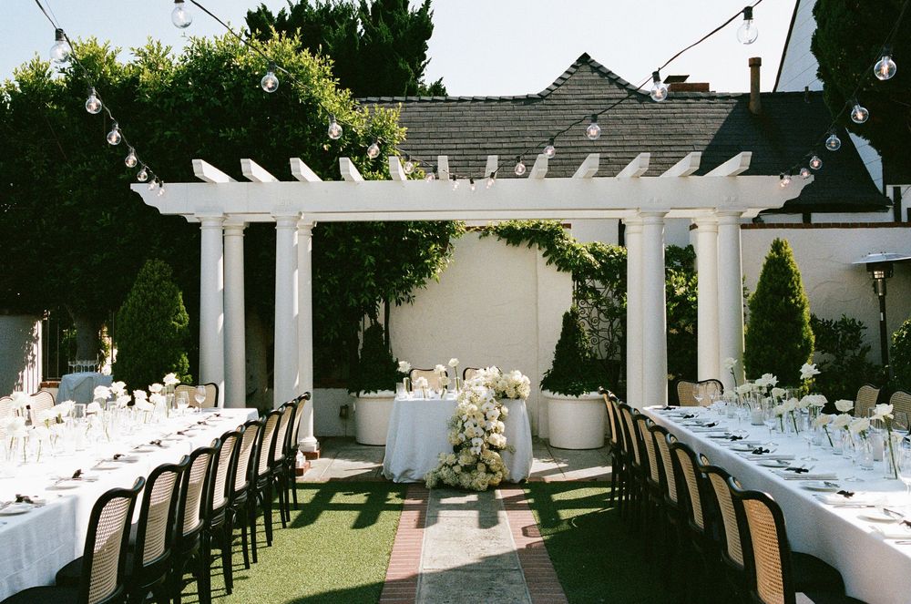 Elegant outdoor wedding reception with white pergola, string lights, long tables with white linens and floral centerpieces.