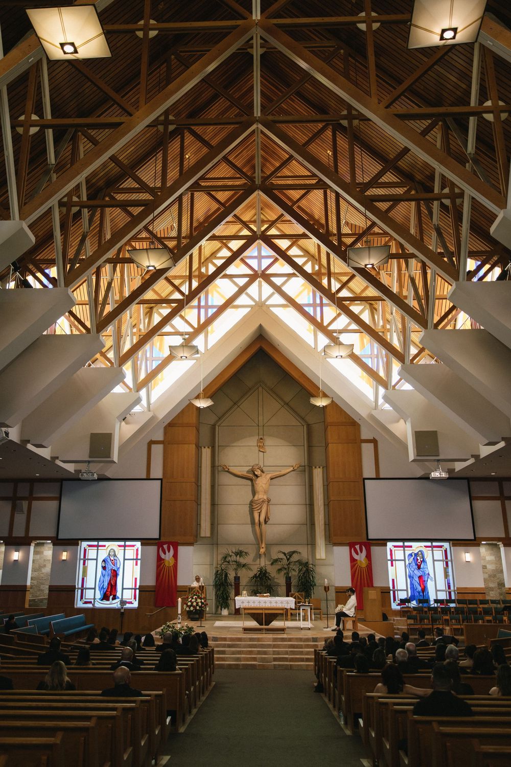 Elegant Catholic church interior with vaulted wooden ceiling, crucifix, stained glass windows, and wedding guests seated in pews.