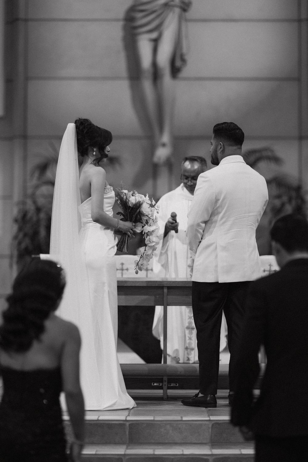Black and white photo of bride and groom exchanging vows at church altar with officiant during wedding ceremony.