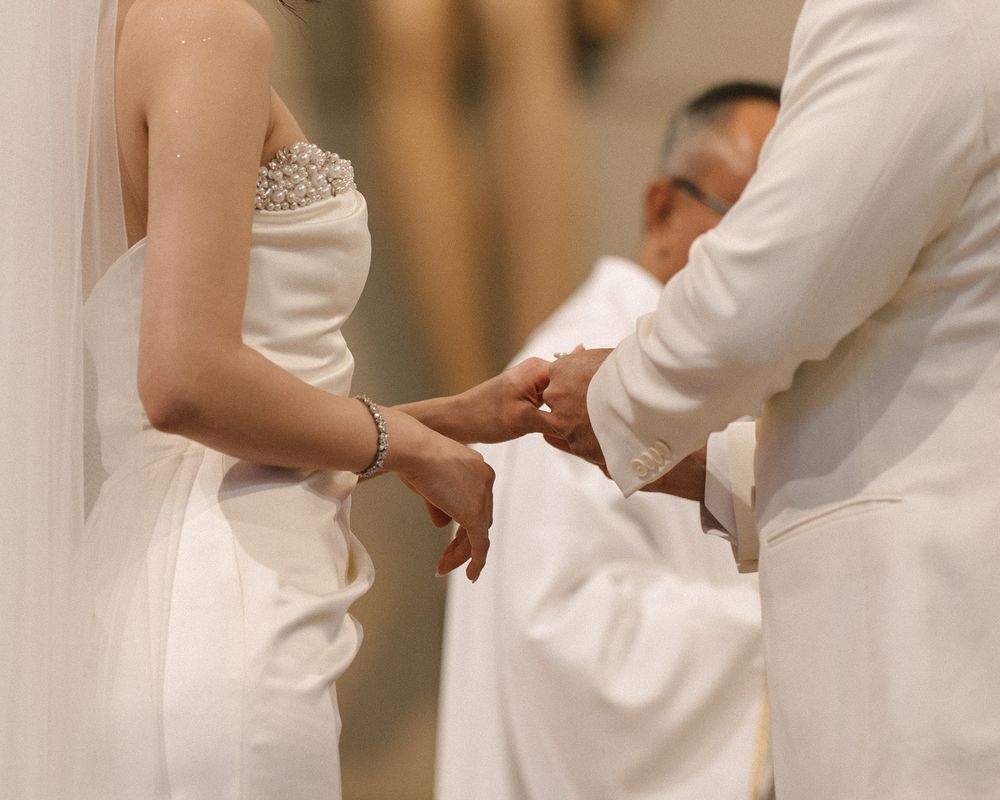 Groom placing wedding ring on bride's finger during intimate church ceremony, bride wearing beaded gown.