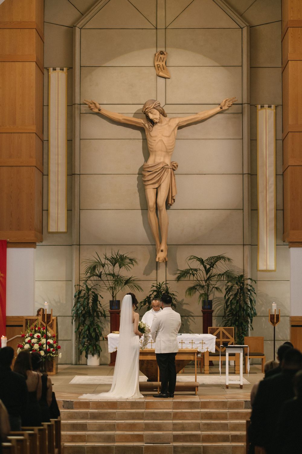 Bride and groom kneeling at altar beneath large crucifix during Catholic wedding ceremony in church.