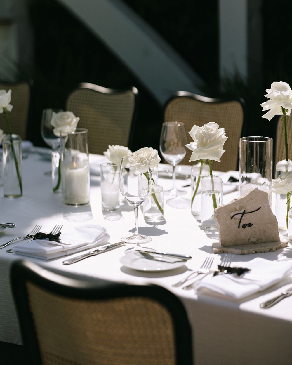 Wedding reception table with white roses in bud vases, wine glasses, candles, and stone table number.