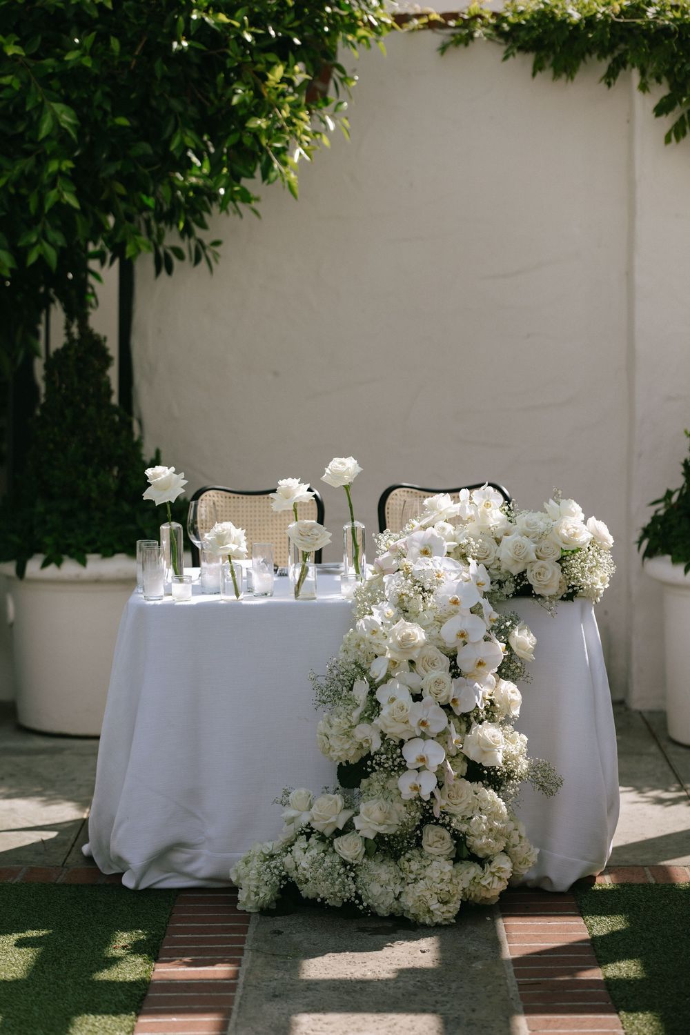 Sweetheart table with lush cascading white floral arrangement of orchids and roses against white wall.
