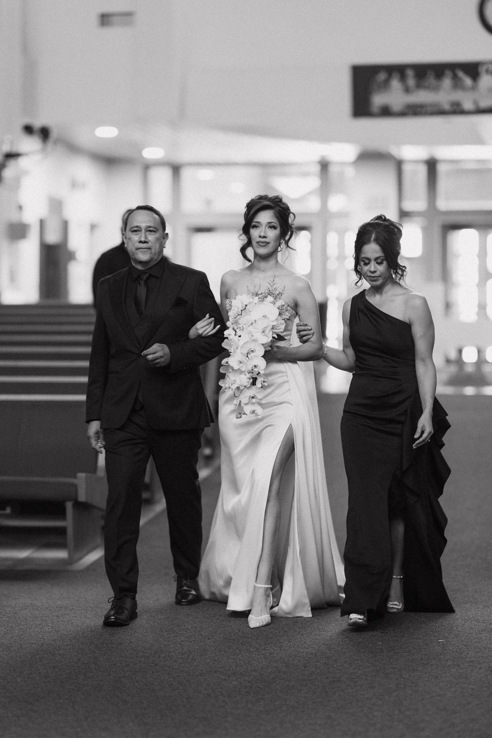 Black and white photo of bride walking down aisle escorted by parents on her wedding day.