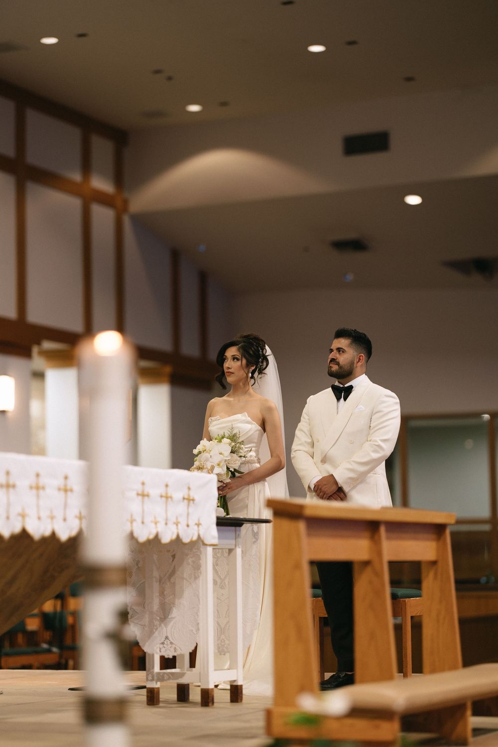 Bride in white strapless gown and groom in white tuxedo standing at church altar during wedding ceremony.