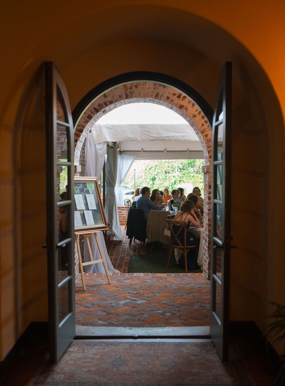 People gather in an arched corridor with brick flooring and white walls leading to a bright outdoor area.
