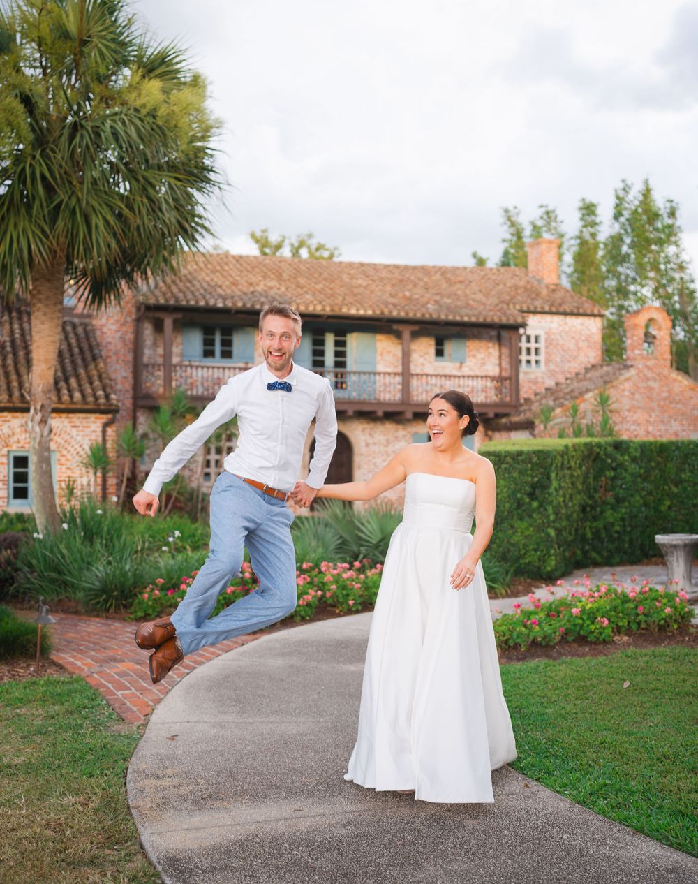 A joyful couple shares a playful moment on a garden path beside a Spanish-style building with palm trees.