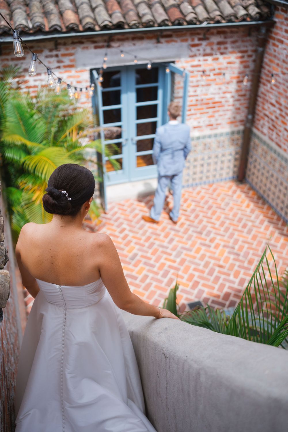 A person in a strapless white dress poses against a rustic brick wall with terra cotta roof tiles and palm plants.