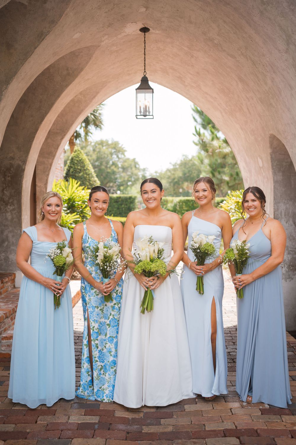 Wedding party stands beneath stone archway wearing light blue bridesmaid dresses and holding yellow floral bouquets.