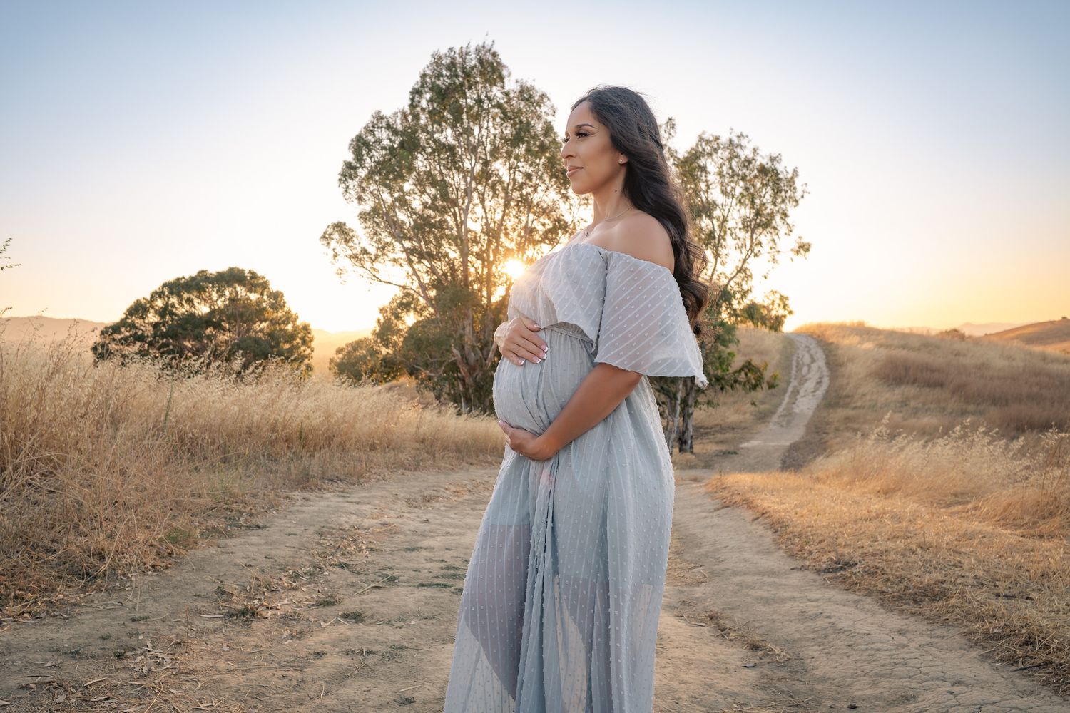 A light blue off-shoulder maternity gown flows gracefully on a dirt path during golden hour sunset with trees in background.