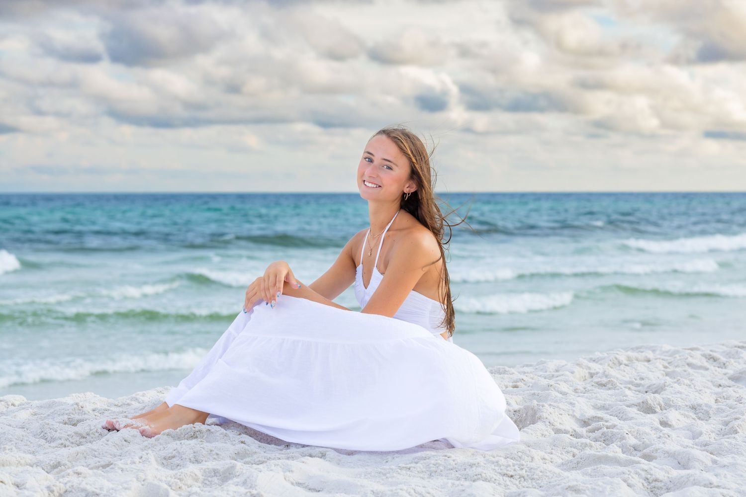 Figure wrapped in white fabric sits peacefully on sandy beach with turquoise ocean waves and cloudy sky backdrop.