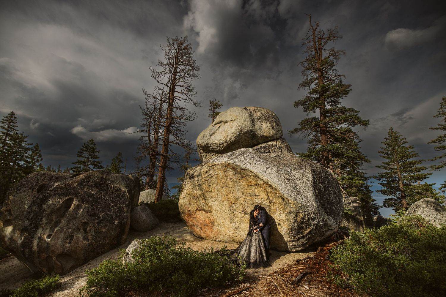 Dramatic stormy sky over rocky mountain terrain with tall pine trees and large boulders.