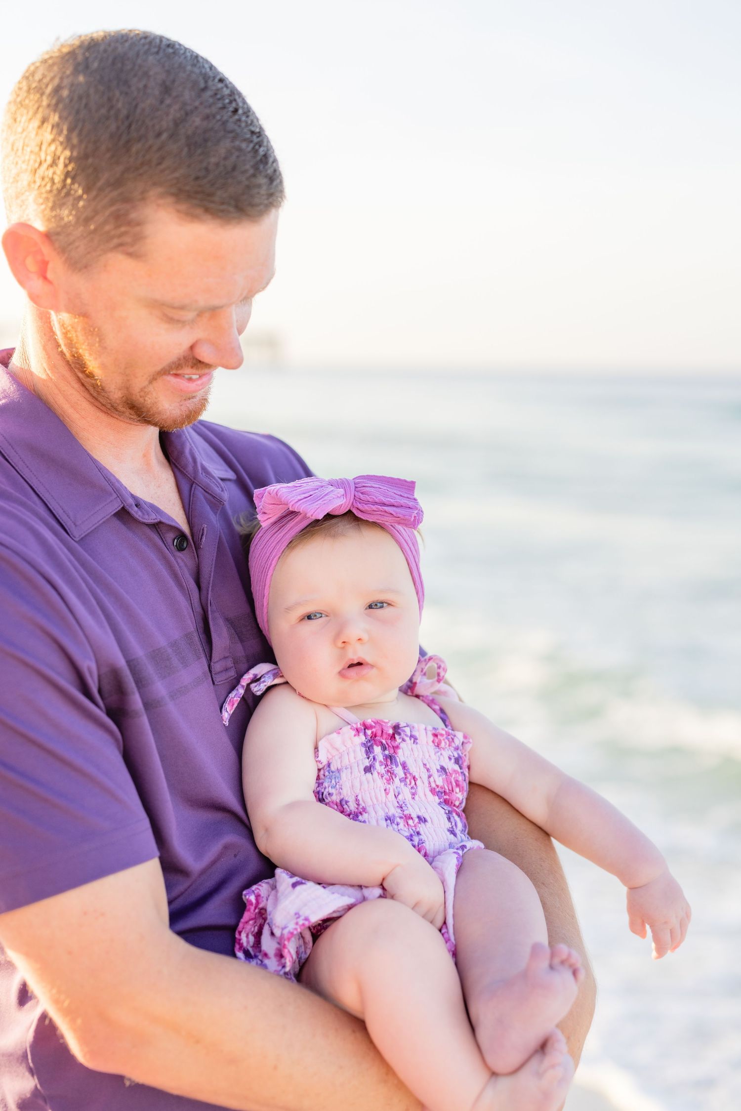 A parent in a purple shirt holds a baby wearing a floral outfit and pink bow at the beach during sunset.