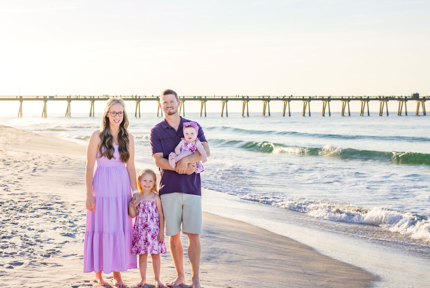 A group of people in coordinated purple outfits pose together on a sunny beach with a wooden pier in the background.