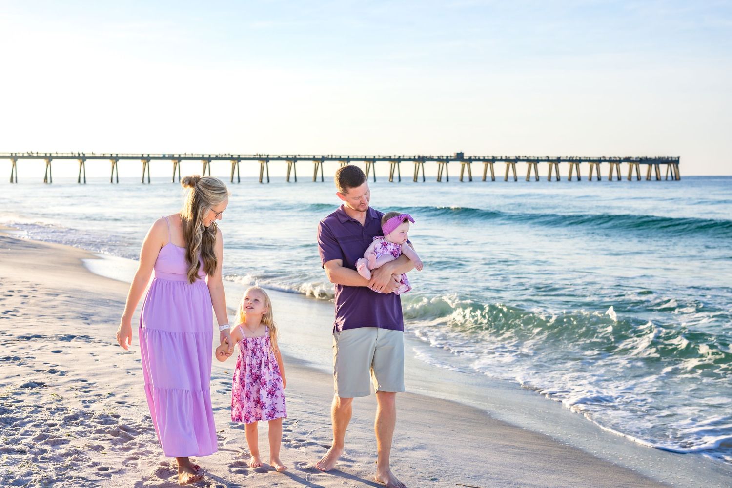 Family walking together along a sandy beach at sunset with a wooden pier in the background.
