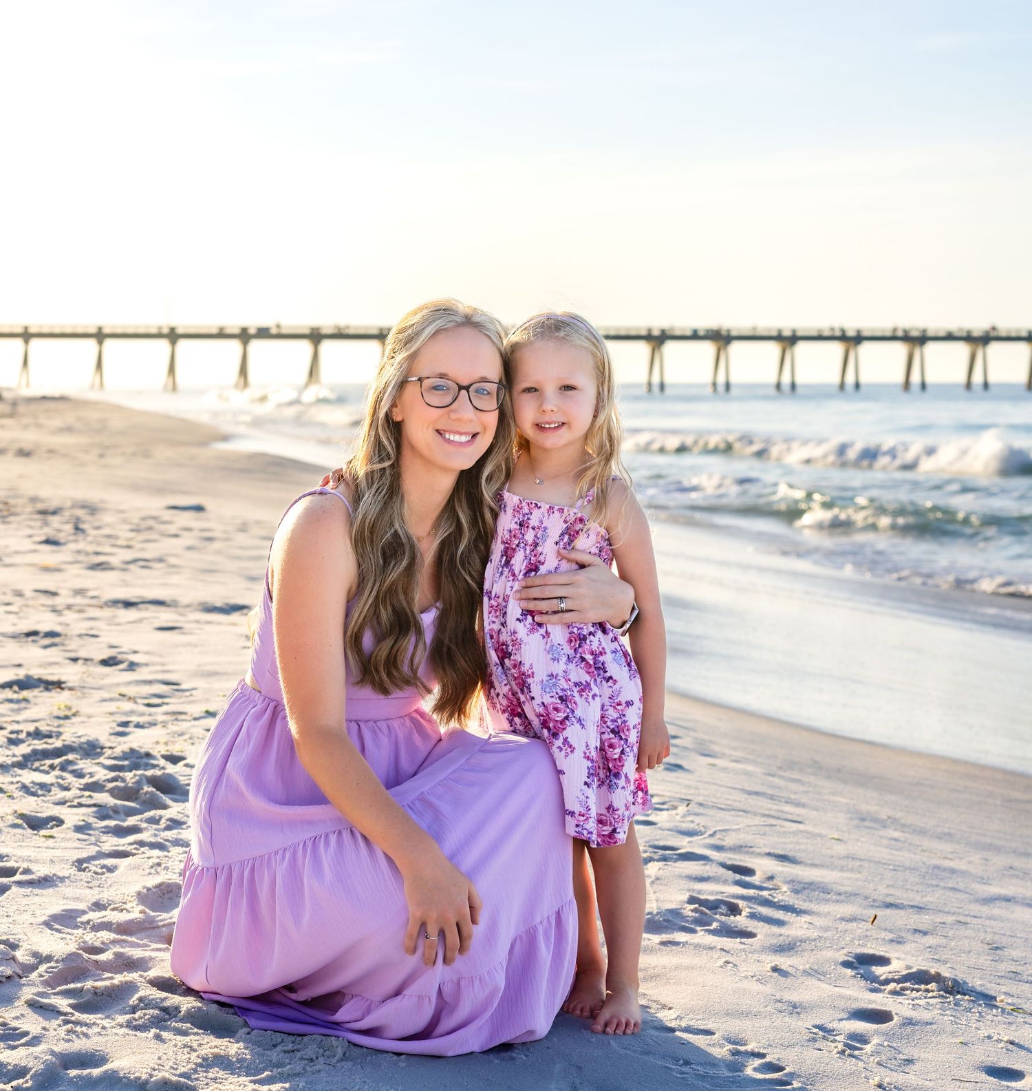 Two people in coordinating purple outfits sit together on a sunny beach with a pier in the background.
