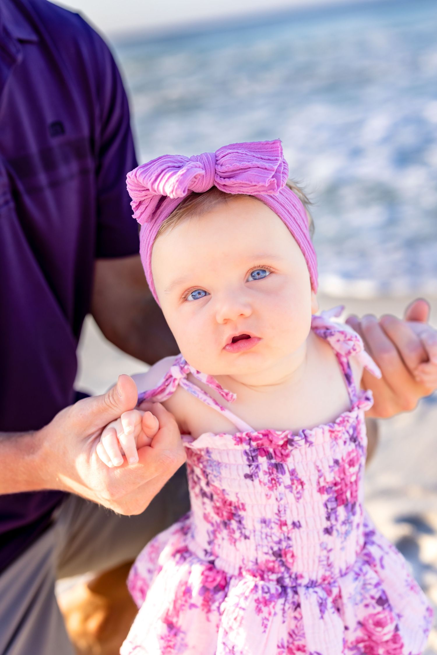 A baby wearing a pink bow headband and floral dress smiles while being held outdoors on a sunny day.