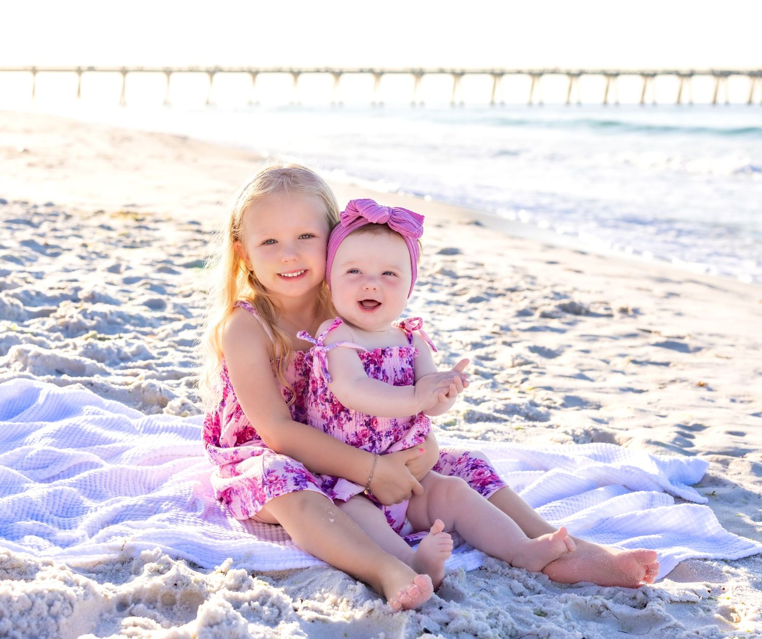 Two children in matching pink floral outfits sit together on a blanket at the beach during sunset.
